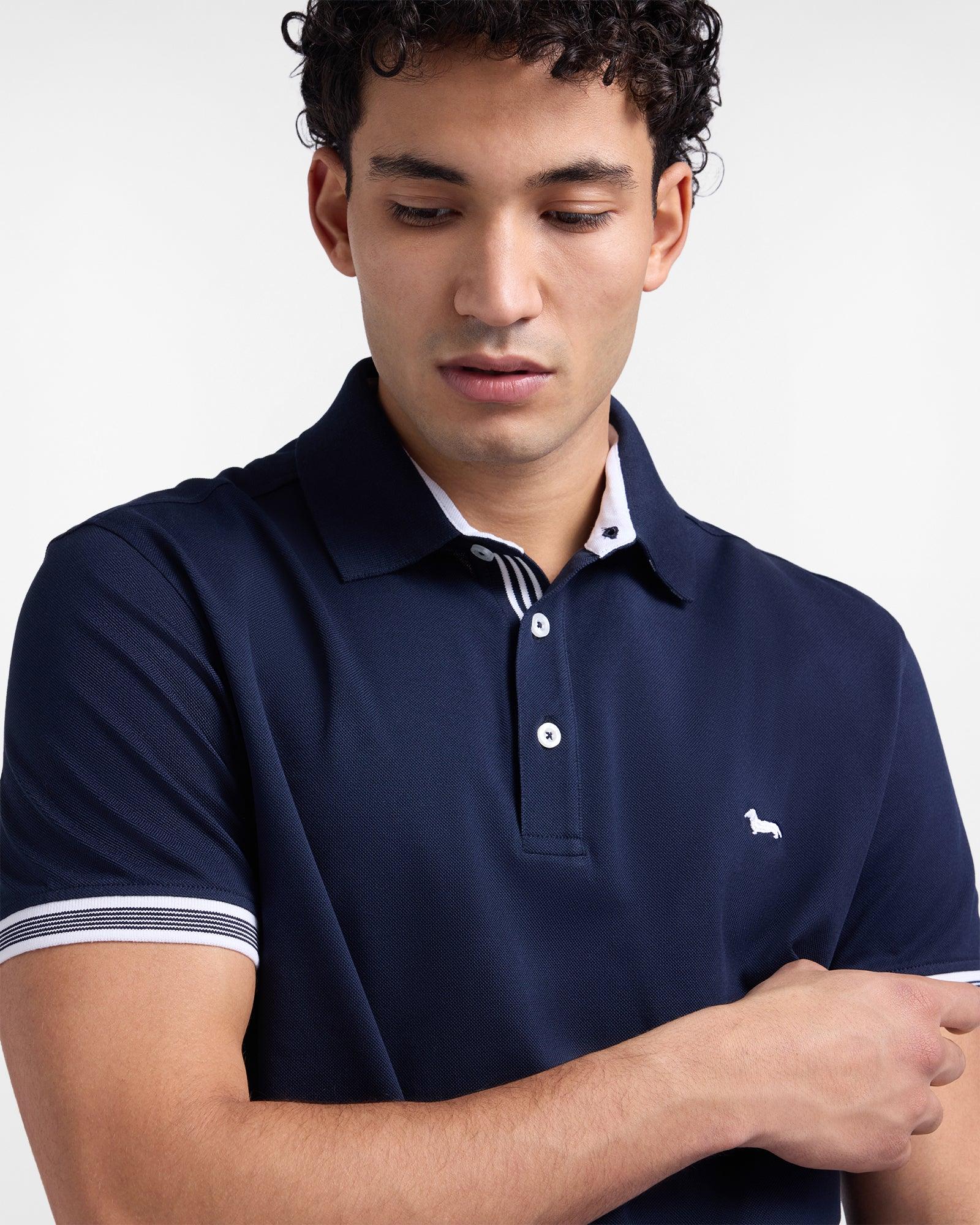 Uomo con polo blu navy a maniche corte, dettagli a righe bianche sul colletto e sulle maniche, logo ricamato sul petto.