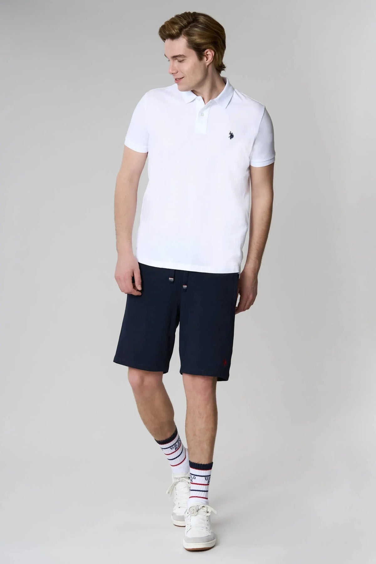 Modello indossa polo bianca e bermuda blu uomo, stile casual estivo, sfondo neutro.