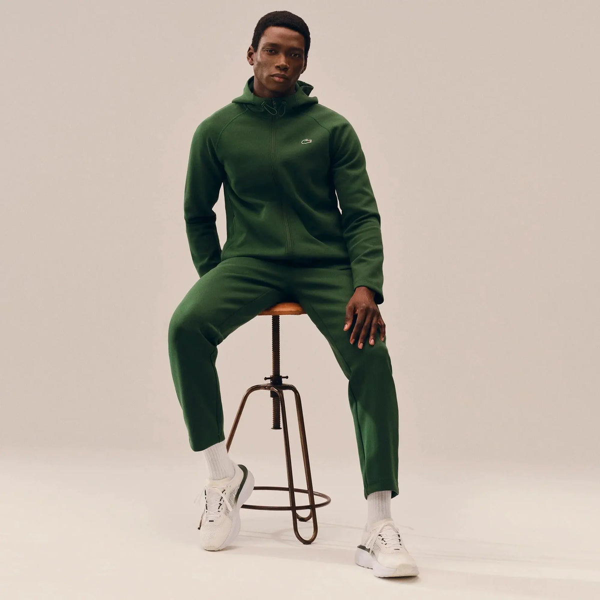 Uomo indossa tuta sportiva verde con cappuccio e sneakers bianche, seduto su sgabello.