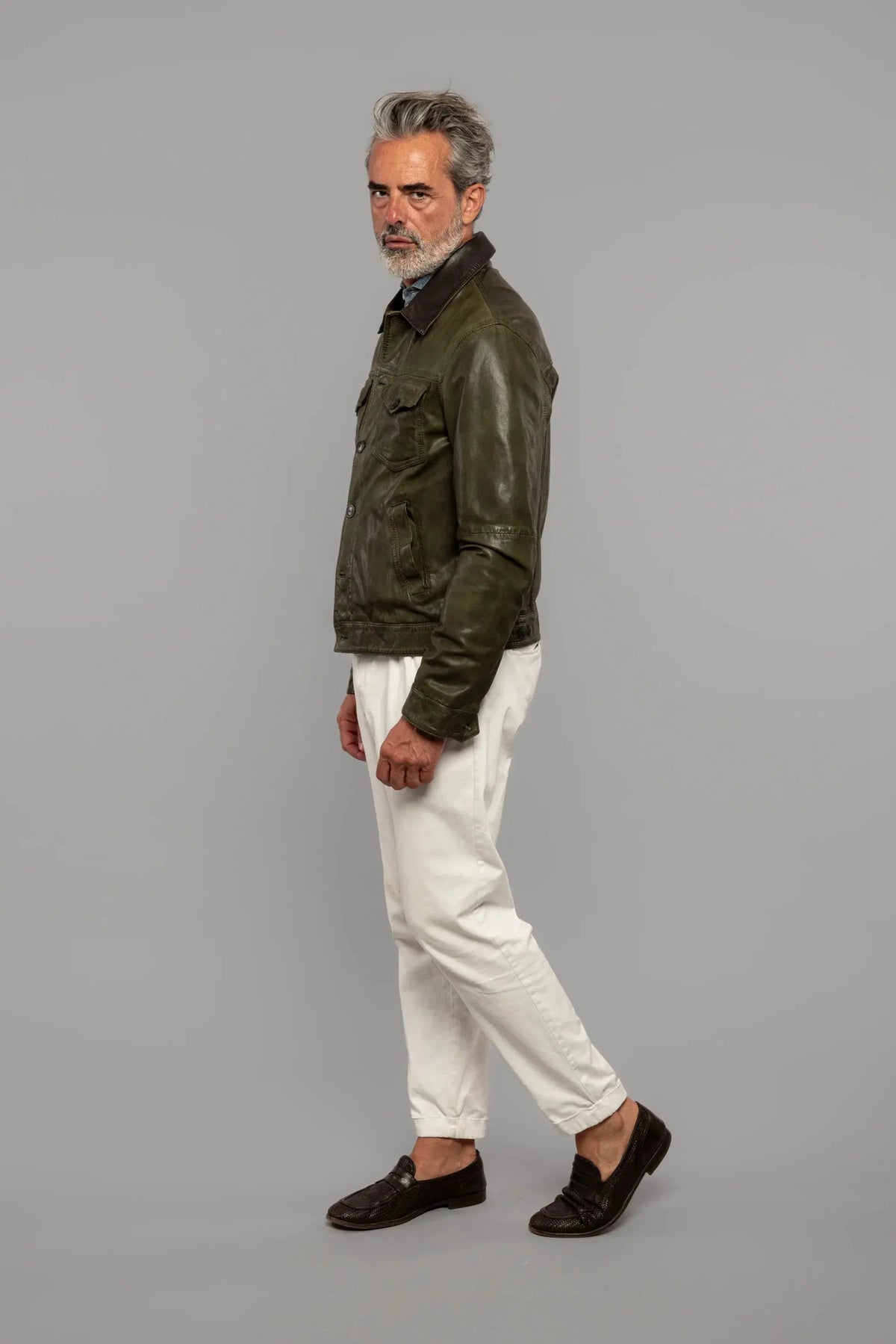 Uomo maturo con giacca in pelle verde, pantaloni bianchi e mocassini neri su sfondo neutro