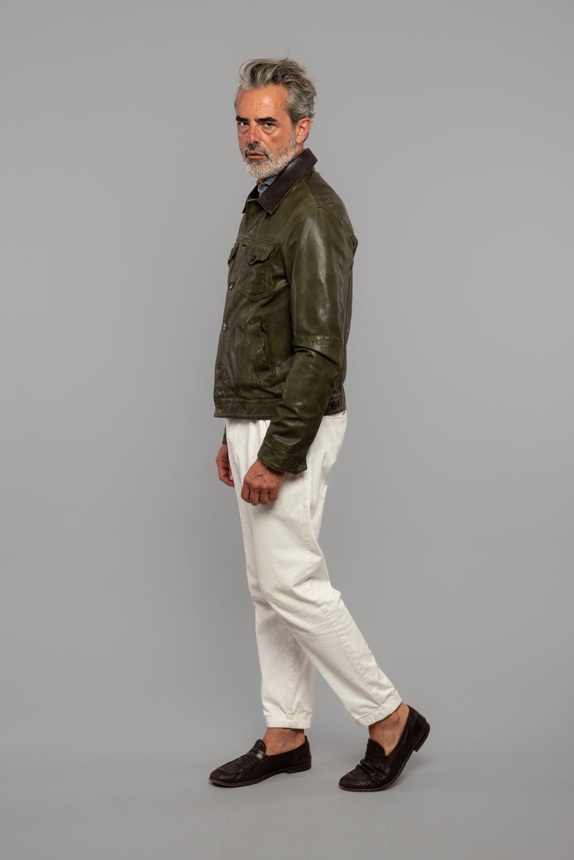Uomo maturo con giacca in pelle verde, pantaloni bianchi e mocassini neri su sfondo neutro