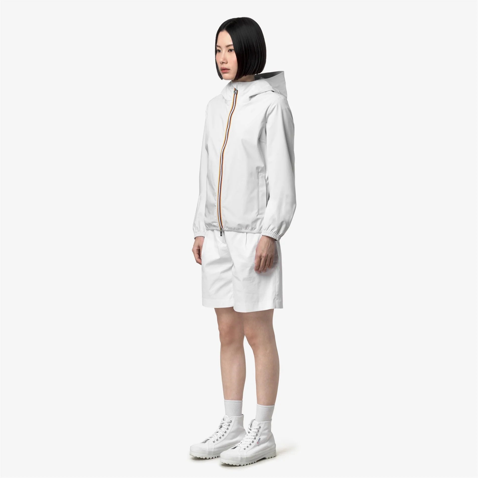 Donna con giacca impermeabile bianca, shorts e scarpe bianche su sfondo neutro
