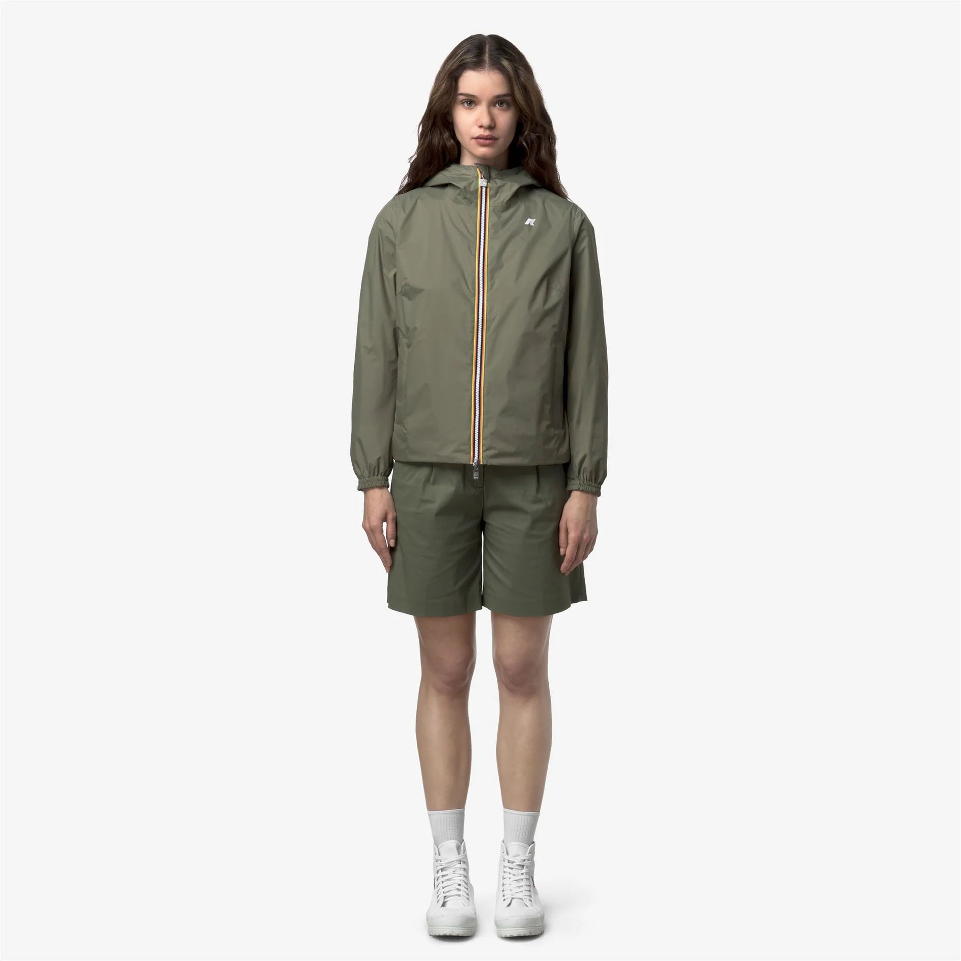 Donna con giacca e shorts verde oliva, stile casual, scarpe bianche, fondo neutro