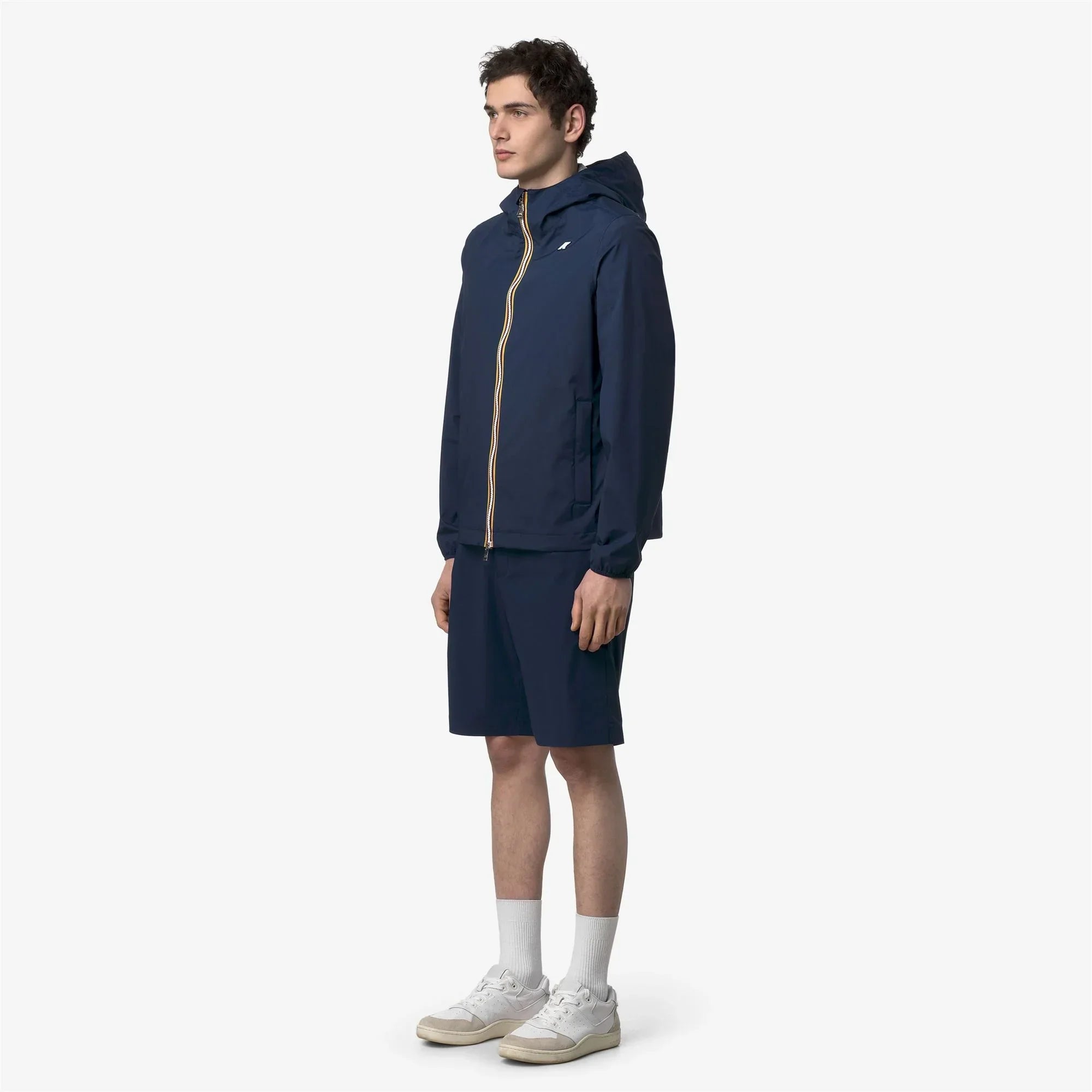 Modello uomo indossa giacca e bermuda blu con cappuccio, stile casual sportivo, su sfondo bianco.