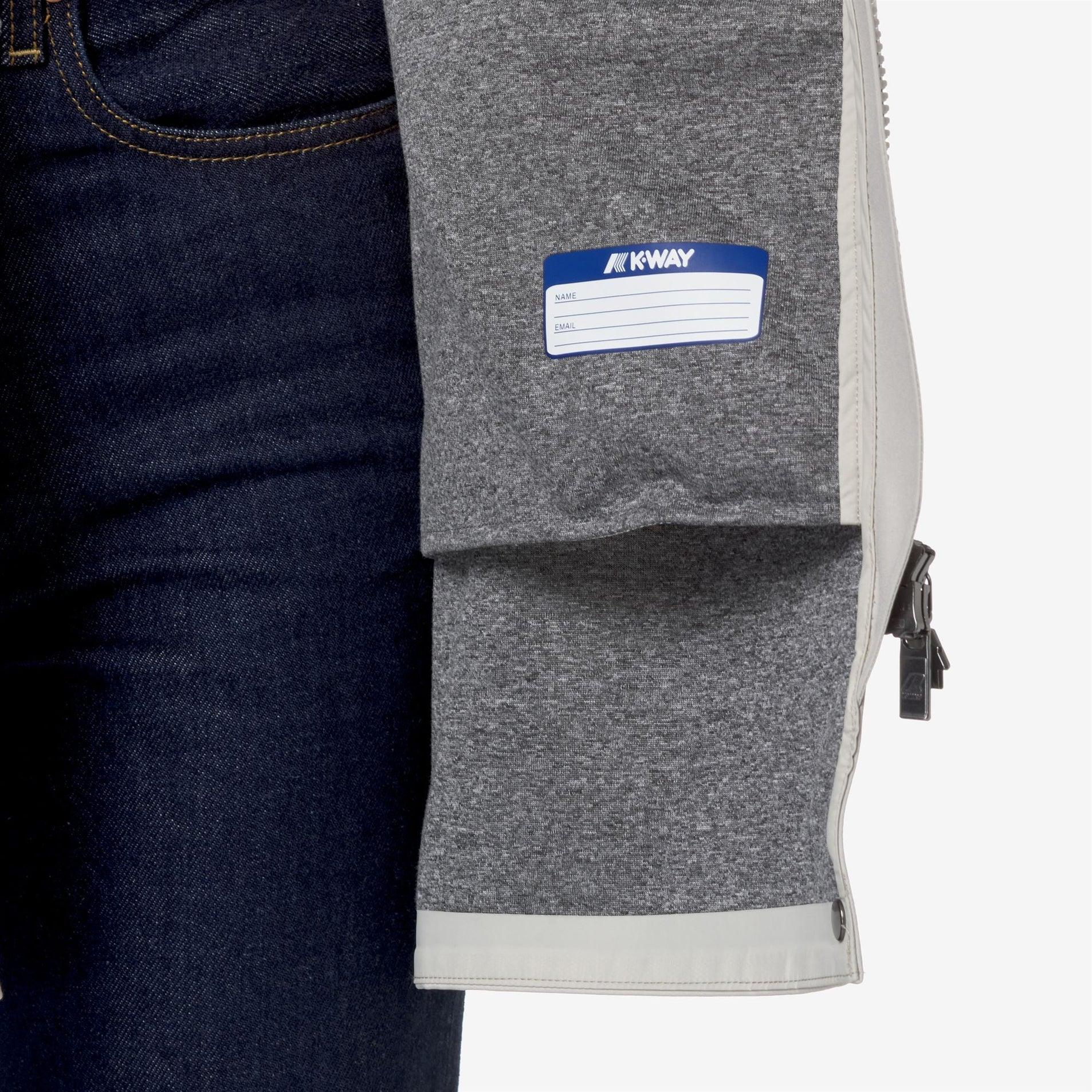 Dettaglio interno giacca grigia K-Way con etichetta personalizzabile e zip, su jeans blu.