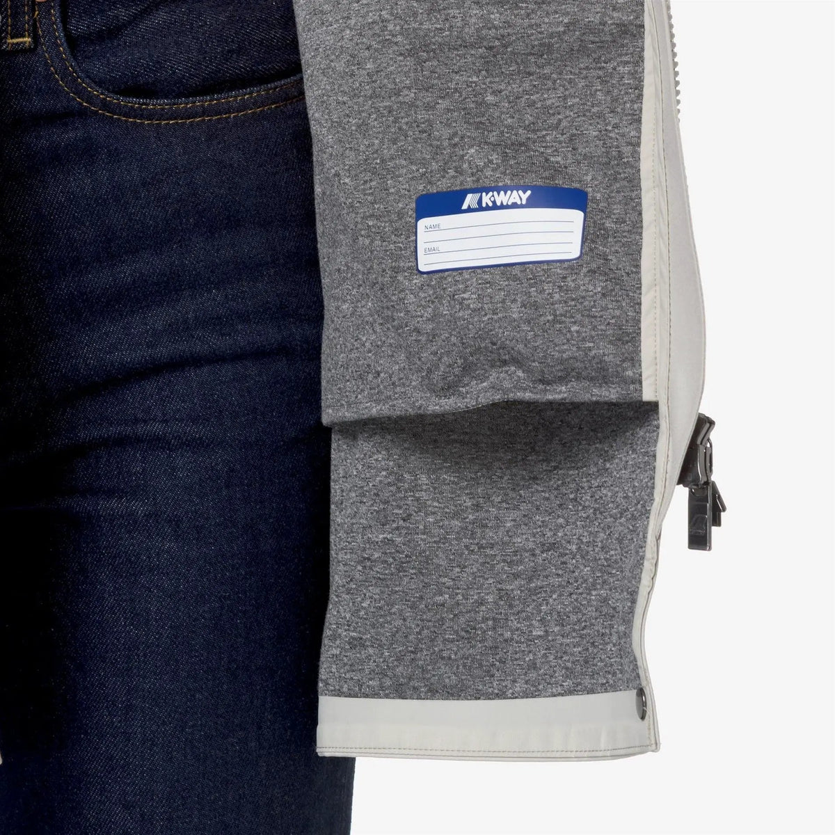Dettaglio interno giacca grigia K-Way con etichetta personalizzabile e zip, su jeans blu.