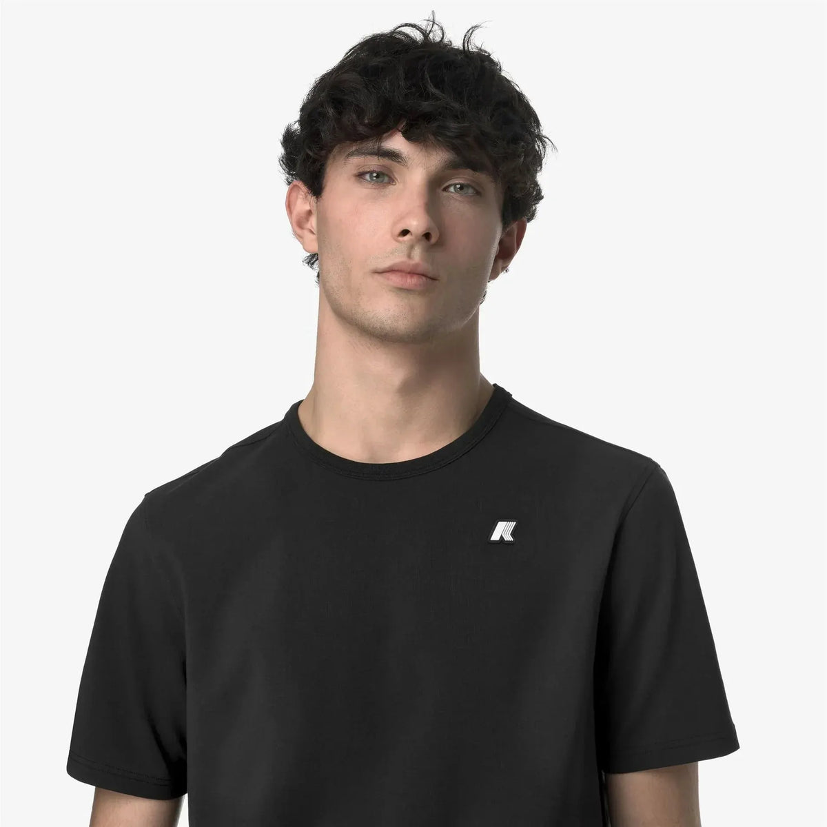 Modello uomo indossa t-shirt nera a manica corta, logo piccolo stampato sul petto, sfondo neutro.