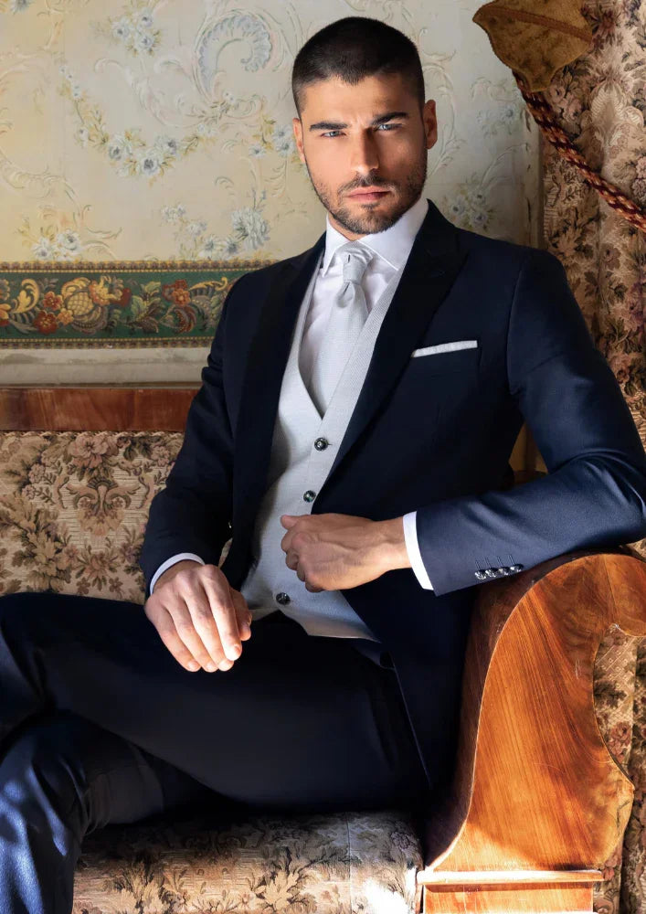 Uomo elegante in abito da cerimonia blu con gilet chiaro, seduto su divano classico, moda uomo.