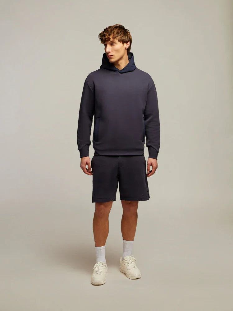 Modello uomo con felpa cappuccio blu e bermuda coordinati, sneakers bianche, su sfondo neutro.
