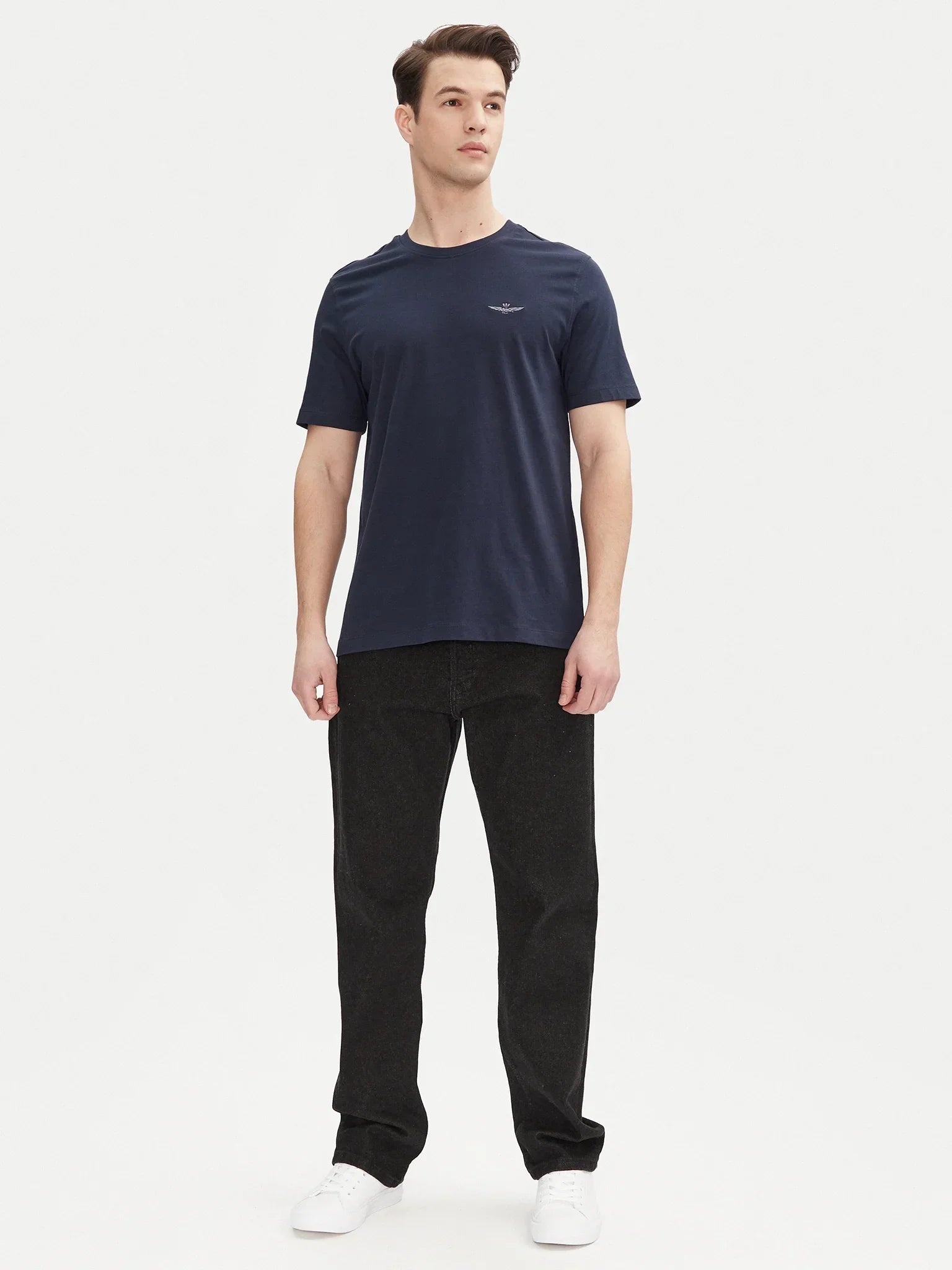Uomo con t-shirt blu navy basic, logo aquila sul petto, jeans neri e sneakers bianche