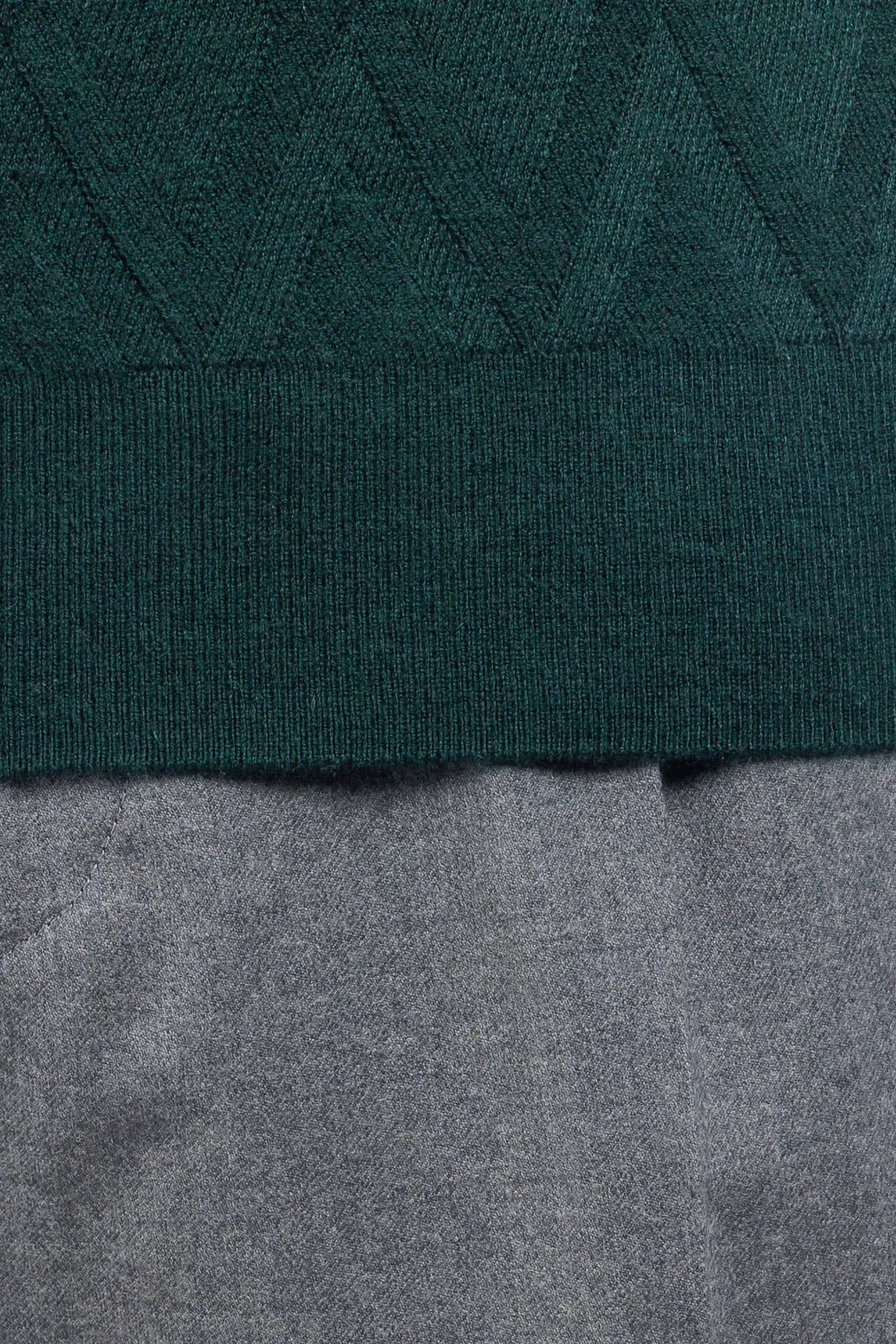 Dettaglio maglia verde bosco lavorata a maglia con motivo geometrico, sopra pantalone grigio.