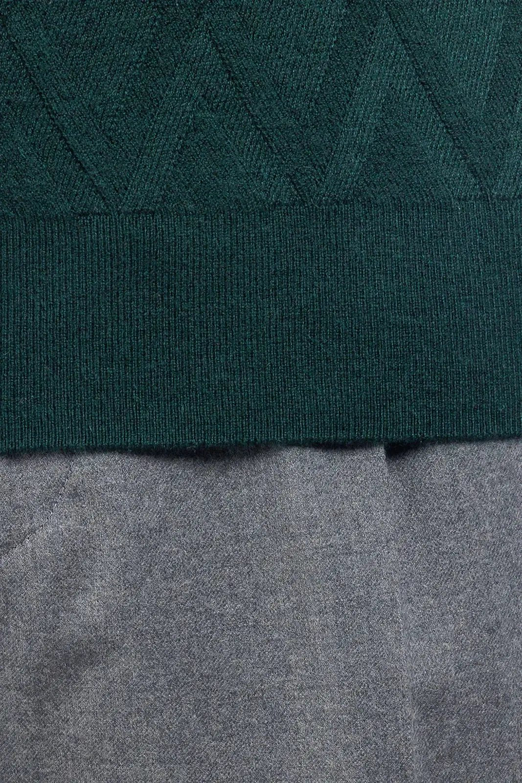 Dettaglio maglia verde bosco lavorata a maglia con motivo geometrico, sopra pantalone grigio.