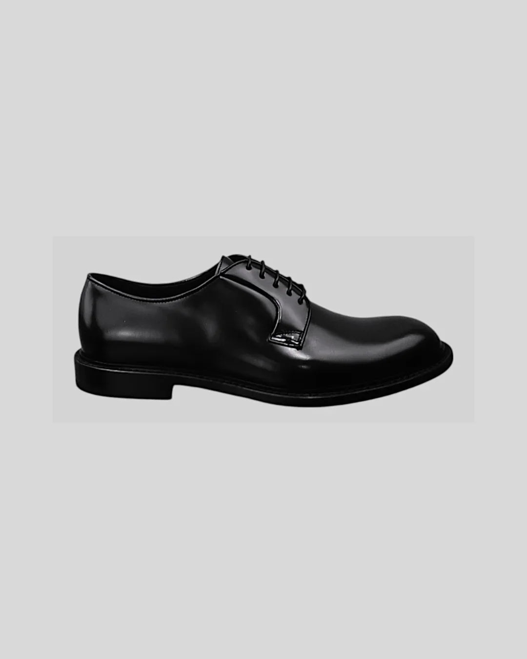 Scarpa derby uomo elegante in pelle nera lucida, con lacci e suola bassa, su sfondo grigio.