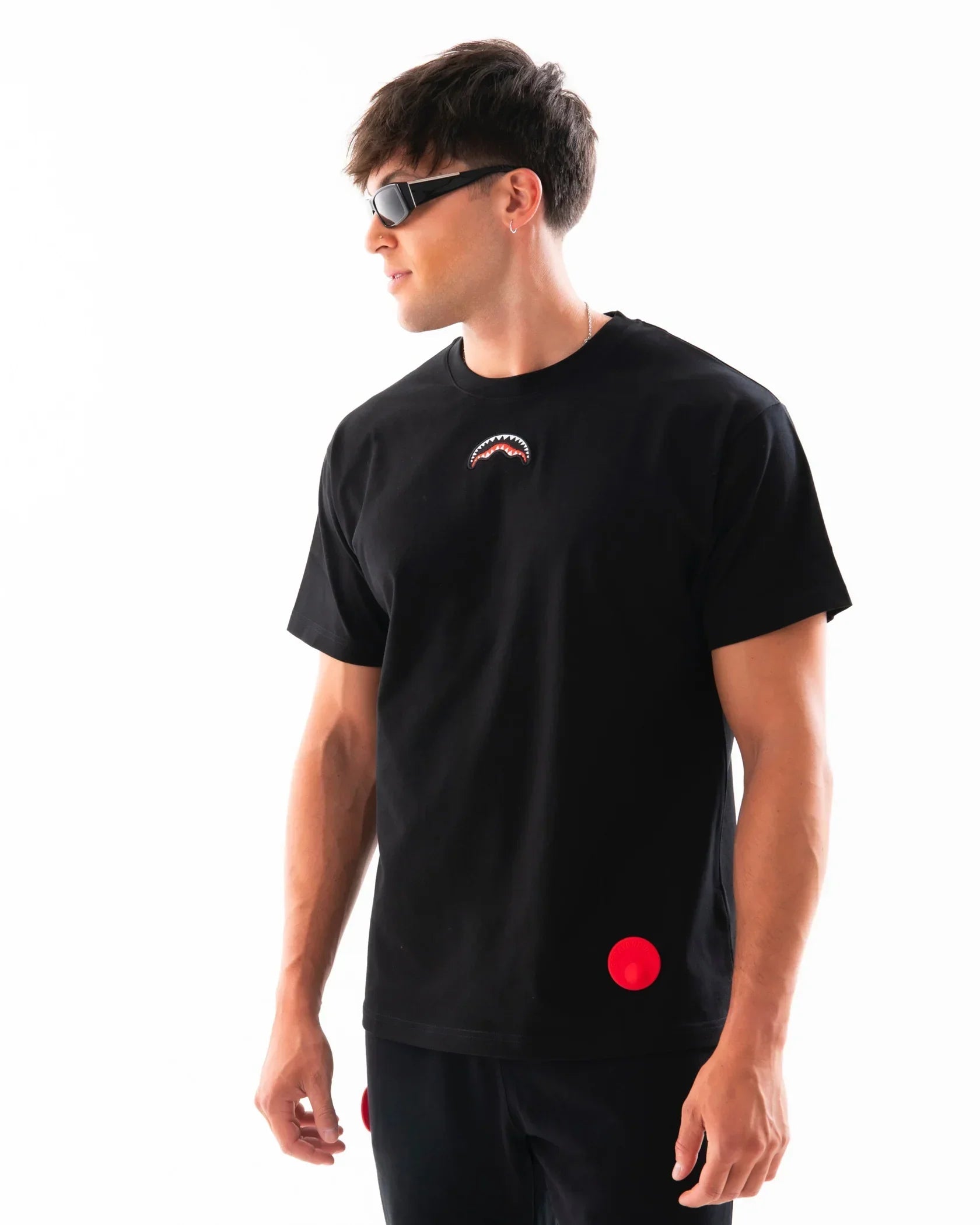 Uomo con t-shirt nera con stampa bocca squalo, pantaloni neri, occhiali da sole moderni
