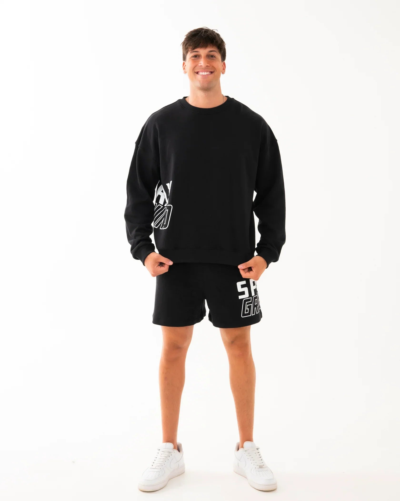 Modello con felpa e shorts neri da uomo, look casual sporty, scarpe bianche, sfondo bianco