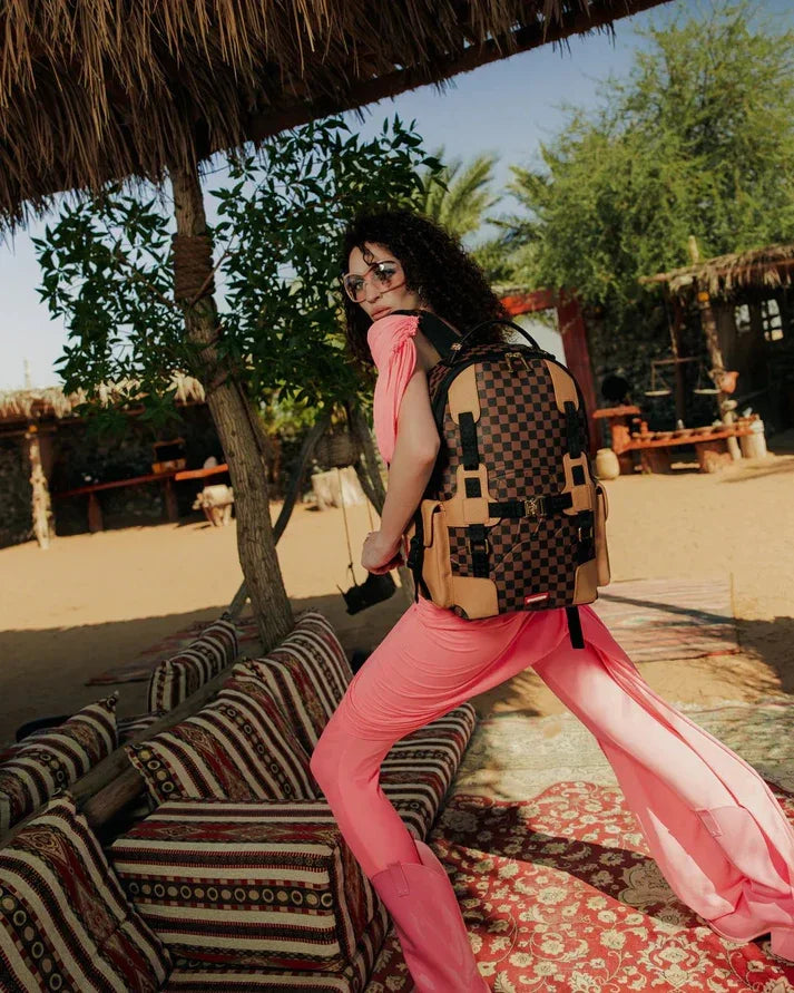 Donna con capelli ricci, occhiali da sole e outfit rosa, zaino a scacchi marrone e nero, in ambiente estivo all'aperto