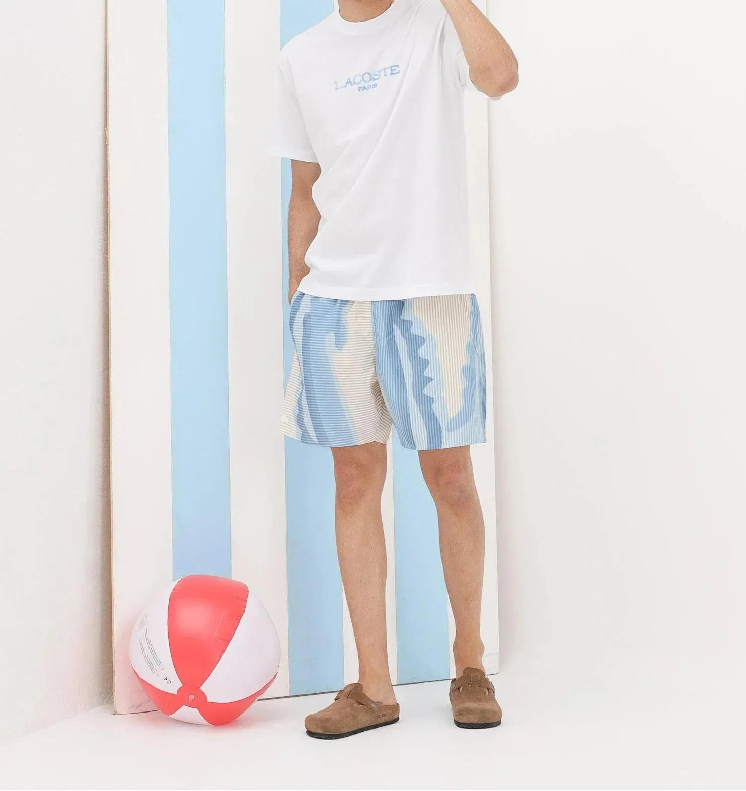 Uomo con t-shirt bianca Lacoste, bermuda blu e beige, sandali estivi e palla da spiaggia.