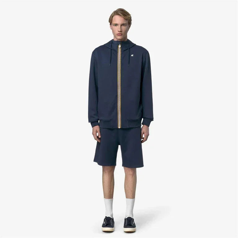 Modello indossa completo sportivo uomo blu con giacca zip e pantaloncini, look casual moderno