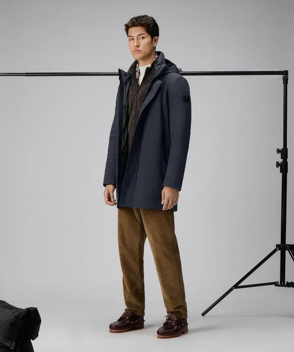 Uomo indossa trench tecnico blu antipioggia, outfit elegante, sfondo neutro.