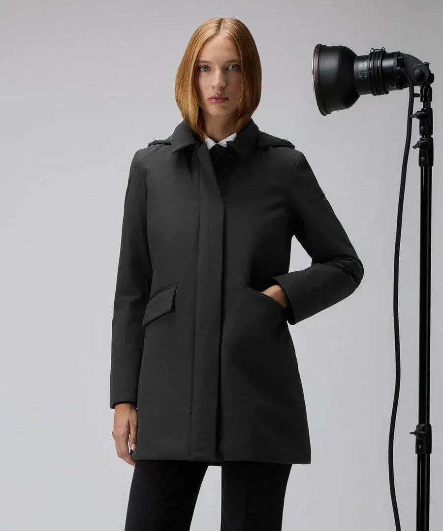 Donna con trench nero dal taglio slim, tasche laterali, look elegante, sfondo neutro.