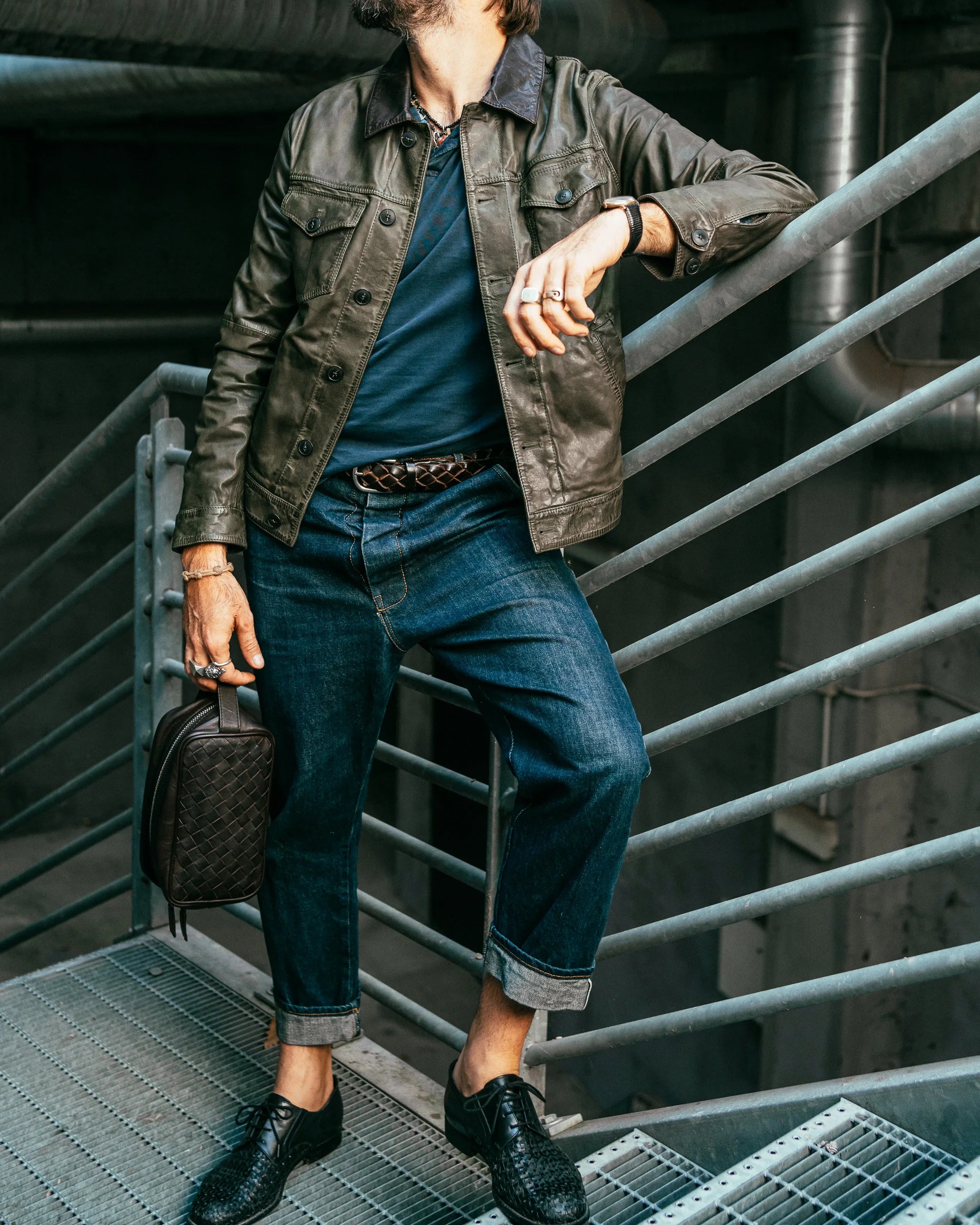Uomo con giacca di pelle verde, jeans risvoltati e scarpe nere intrecciate, stile casual elegante.
