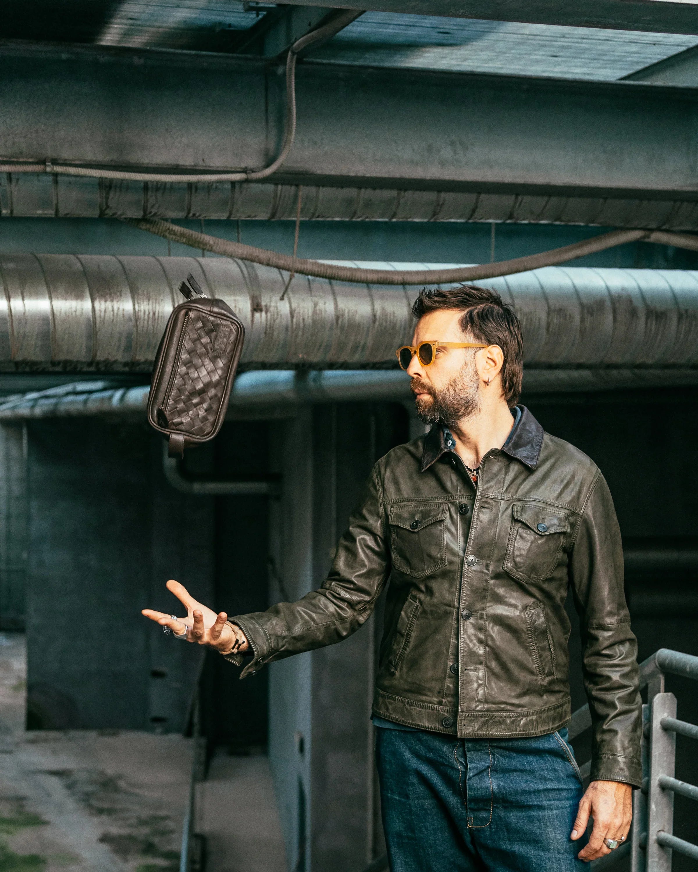 Uomo con giacca in pelle verde, jeans e occhiali da sole gialli in ambiente industriale