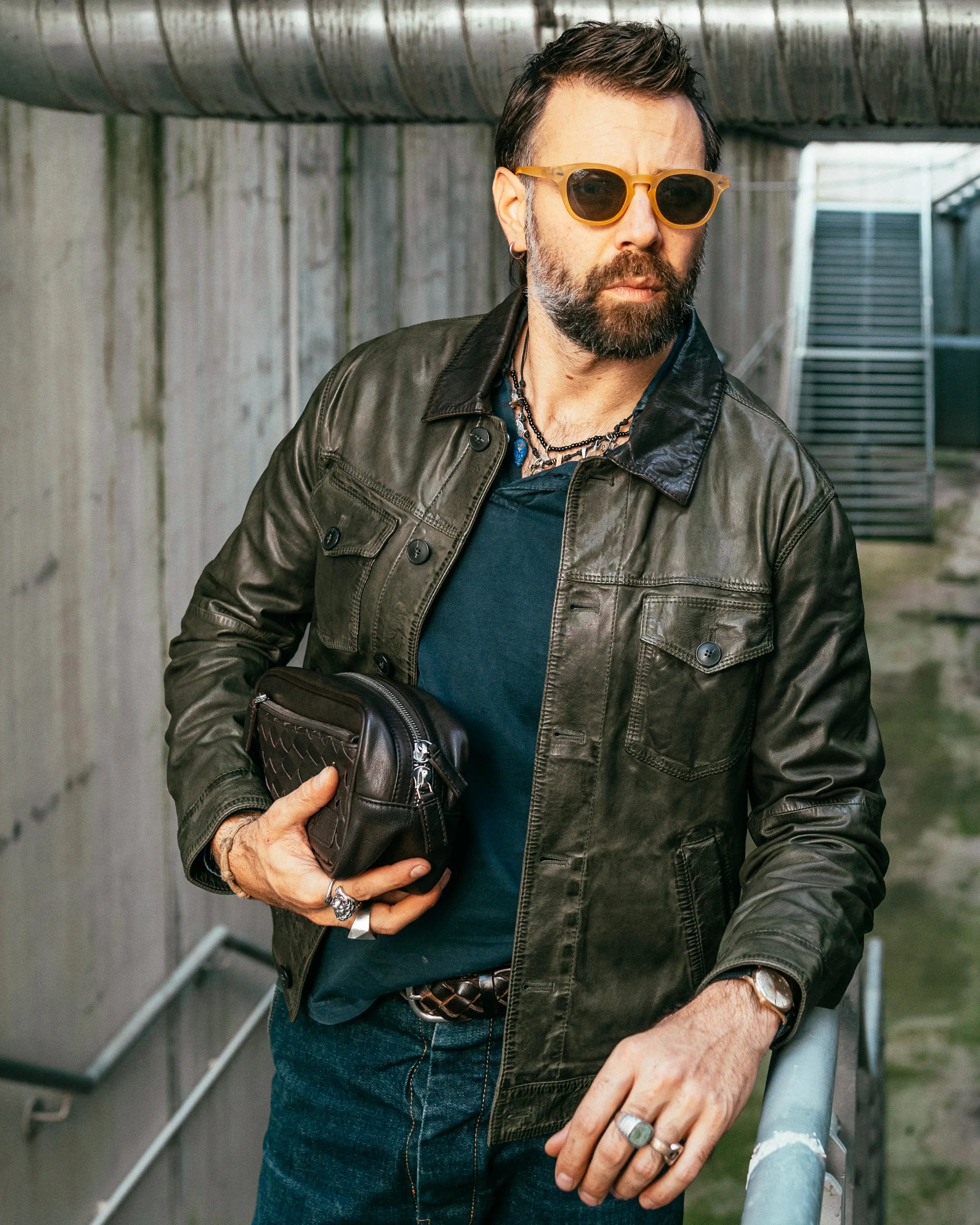 Uomo con giacca di pelle verde, jeans e occhiali da sole, tiene una borsa in mano.