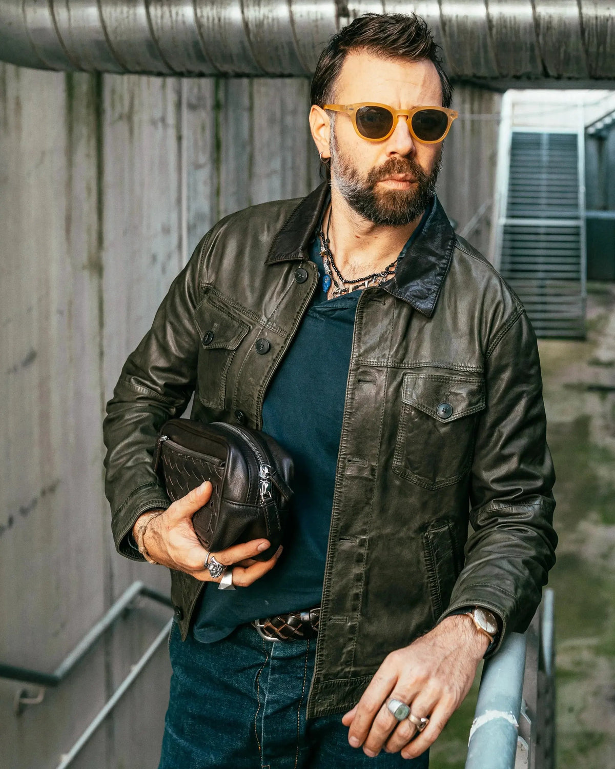 Uomo con giacca di pelle verde, jeans e occhiali da sole, tiene una borsa in mano.