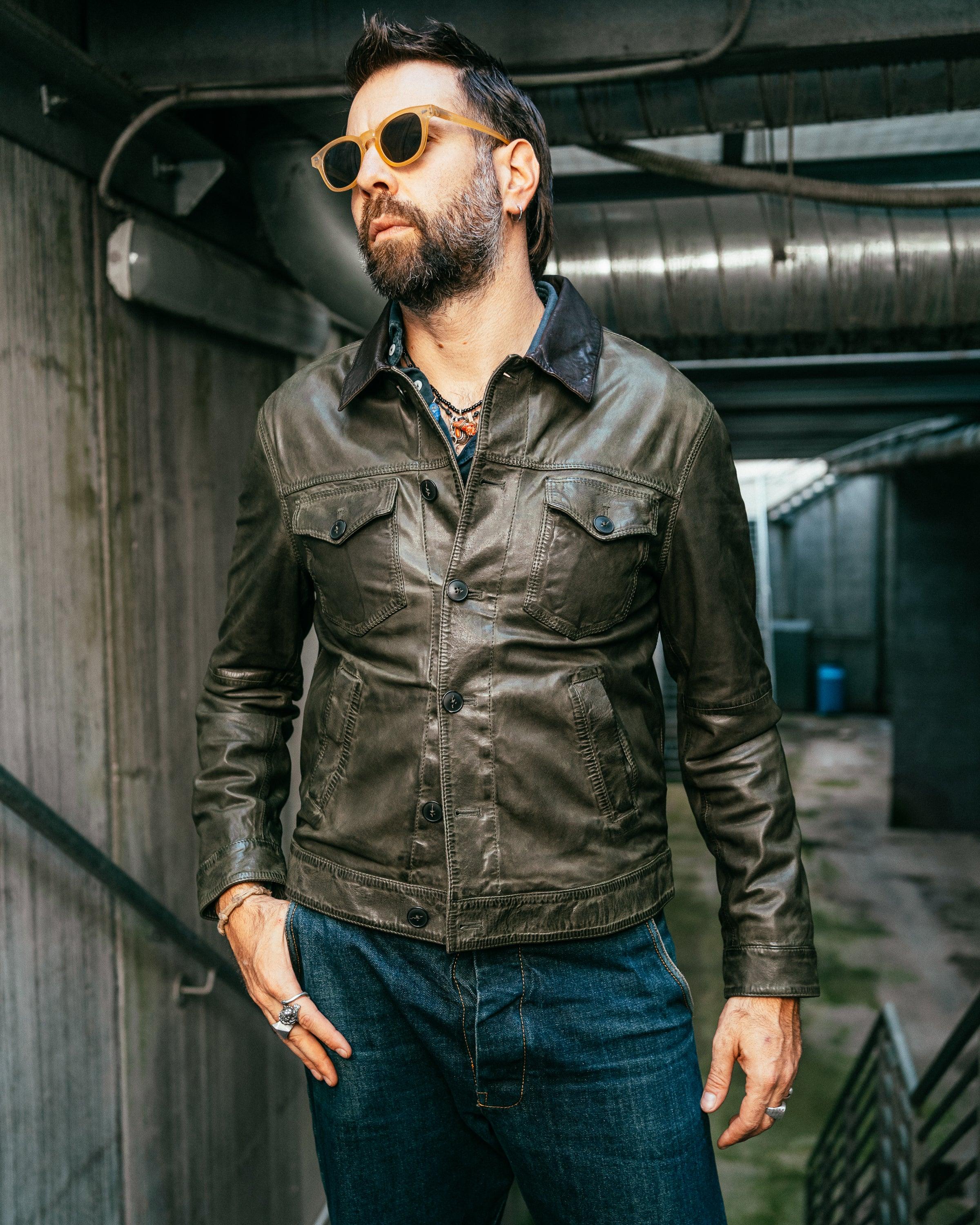 Uomo con giacca in pelle verde oliva, jeans blu e occhiali da sole, stile casual urbano.