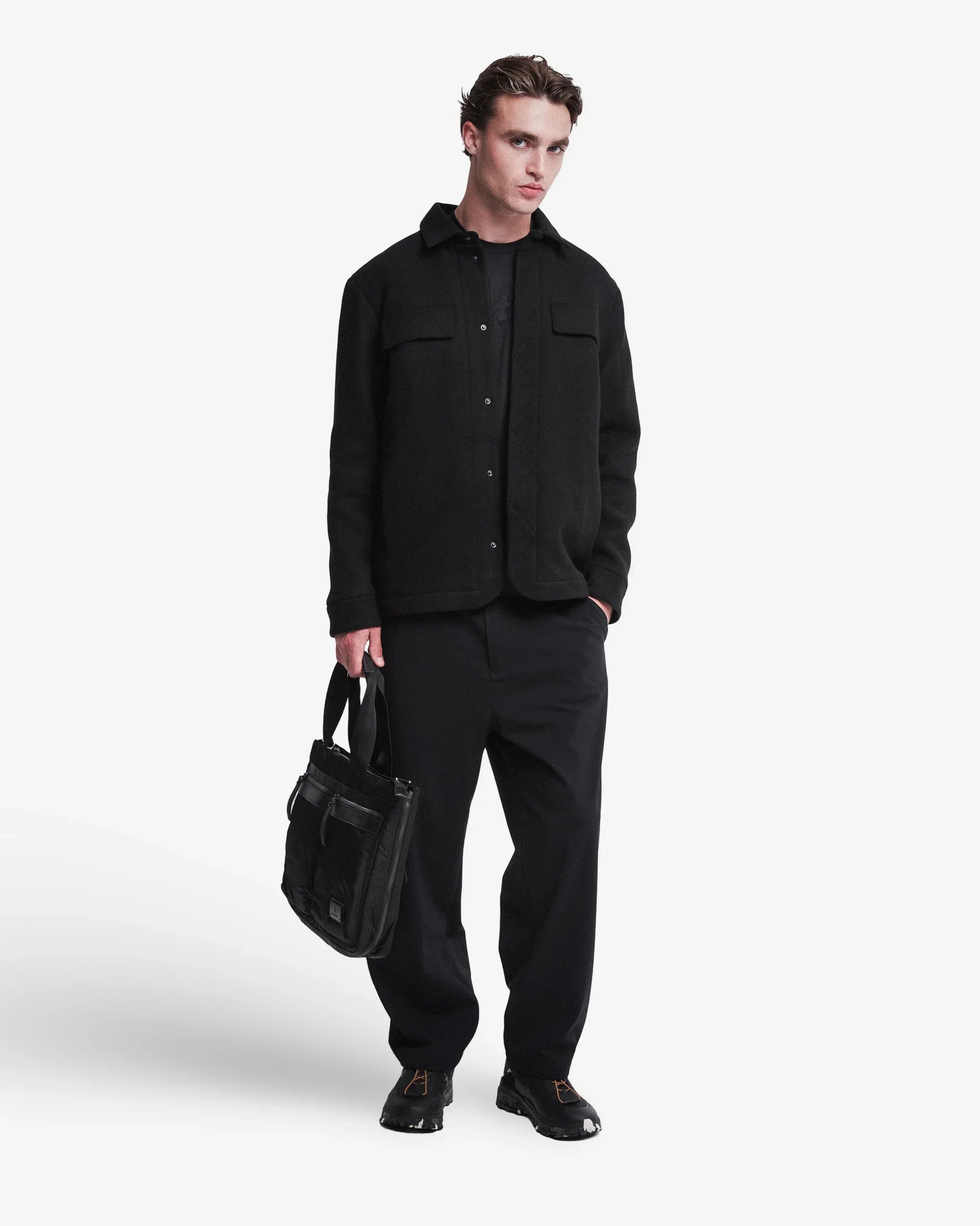 Uomo con giacca nera elegante, pantaloni neri e borsa a mano, su sfondo bianco