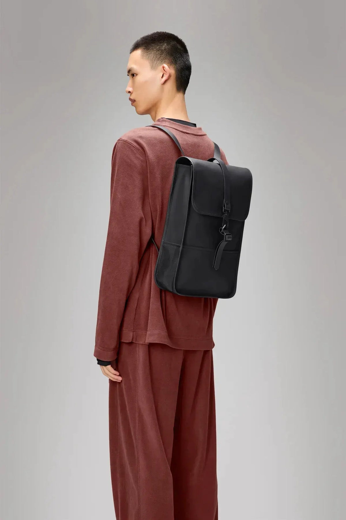 Zaino nero minimalista da uomo indossato su spalle, outfit marrone, sfondo neutro.