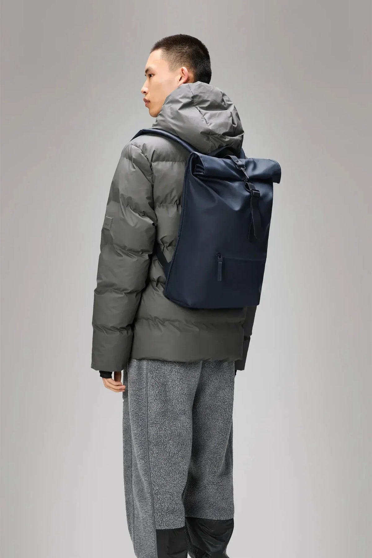 Modello con zaino blu navy roll-top e giacca imbottita grigia, look moderno uomo.