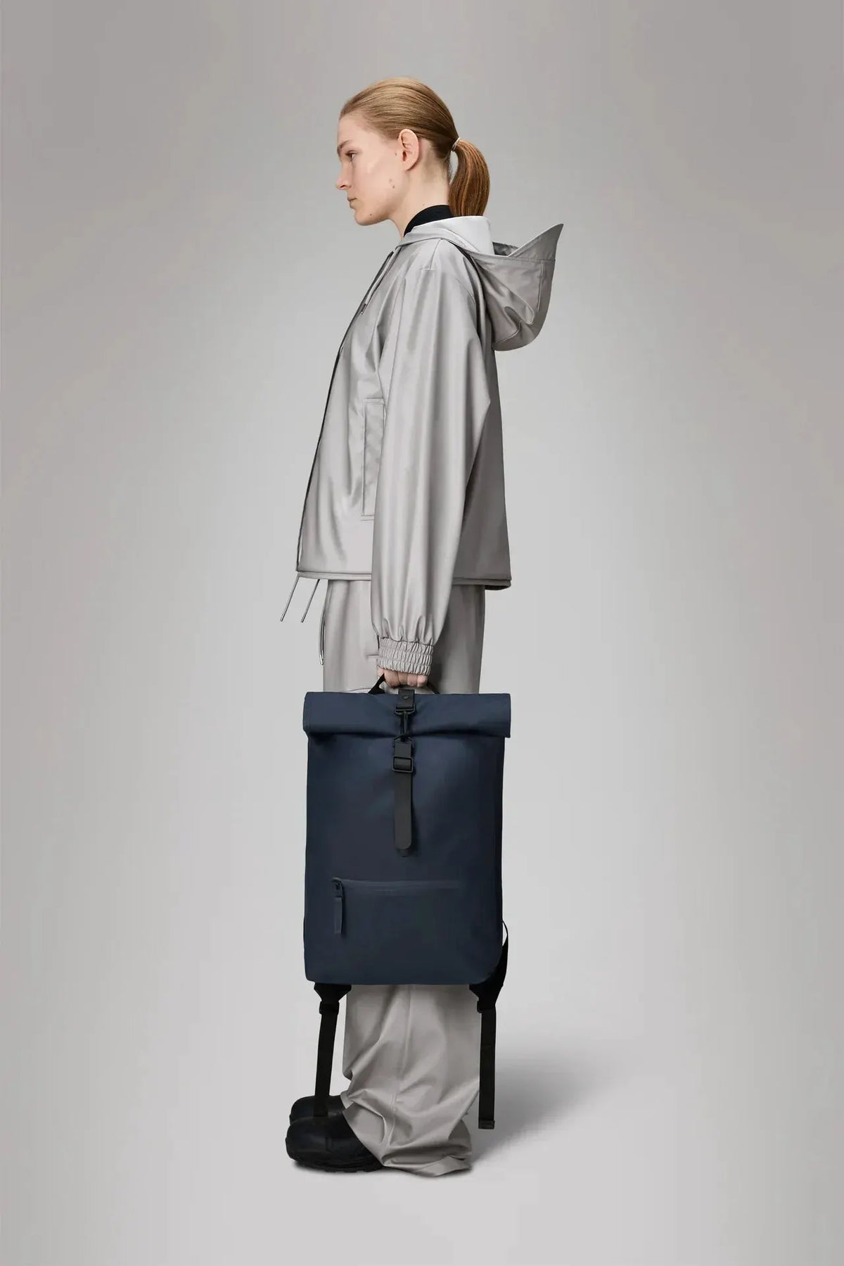 Zaino rolltop blu in materiale impermeabile tenuto da donna con outfit grigio, vista laterale