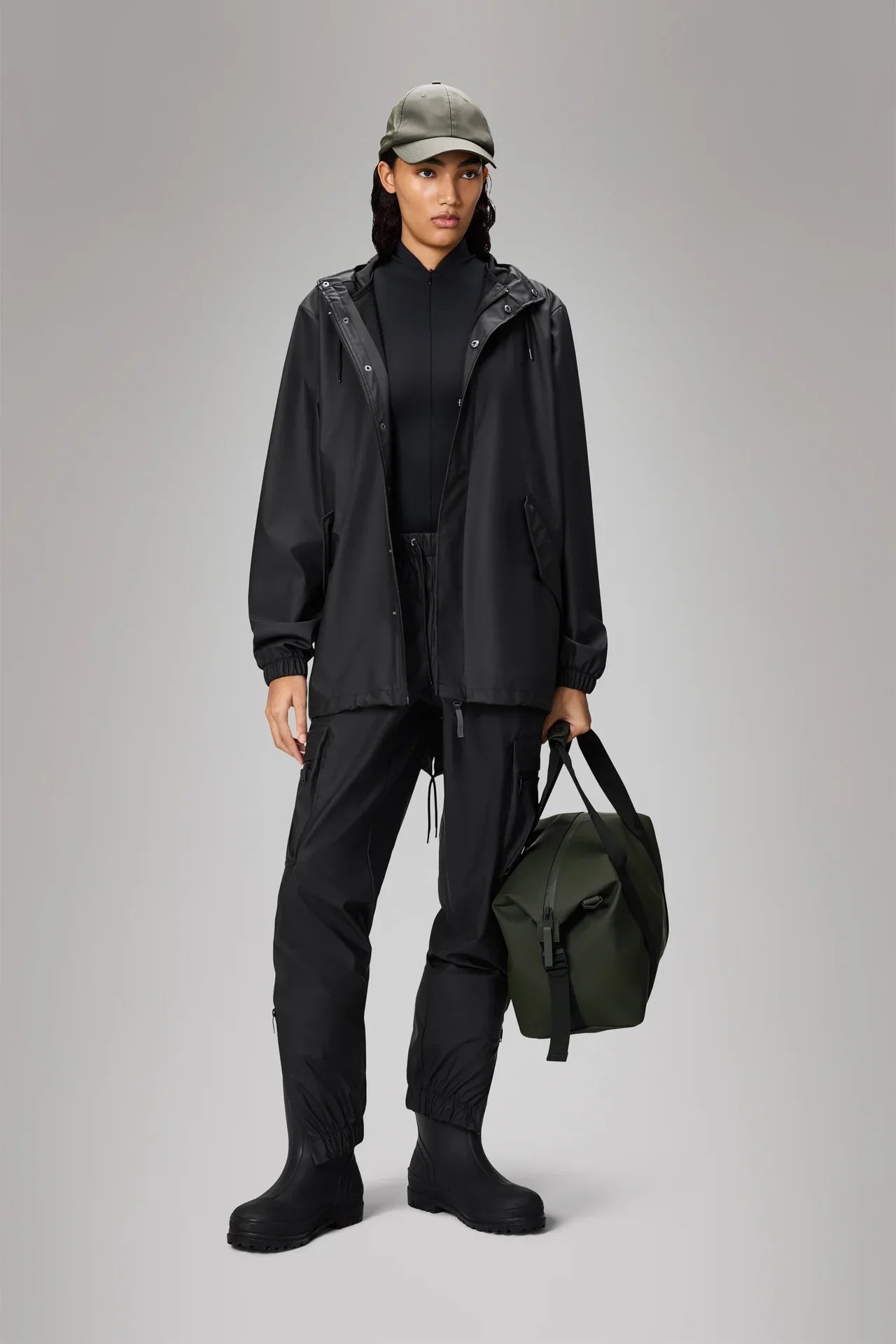 Donna con giacca impermeabile nera, pantaloni tecnici e borsa verde, stile outdoor