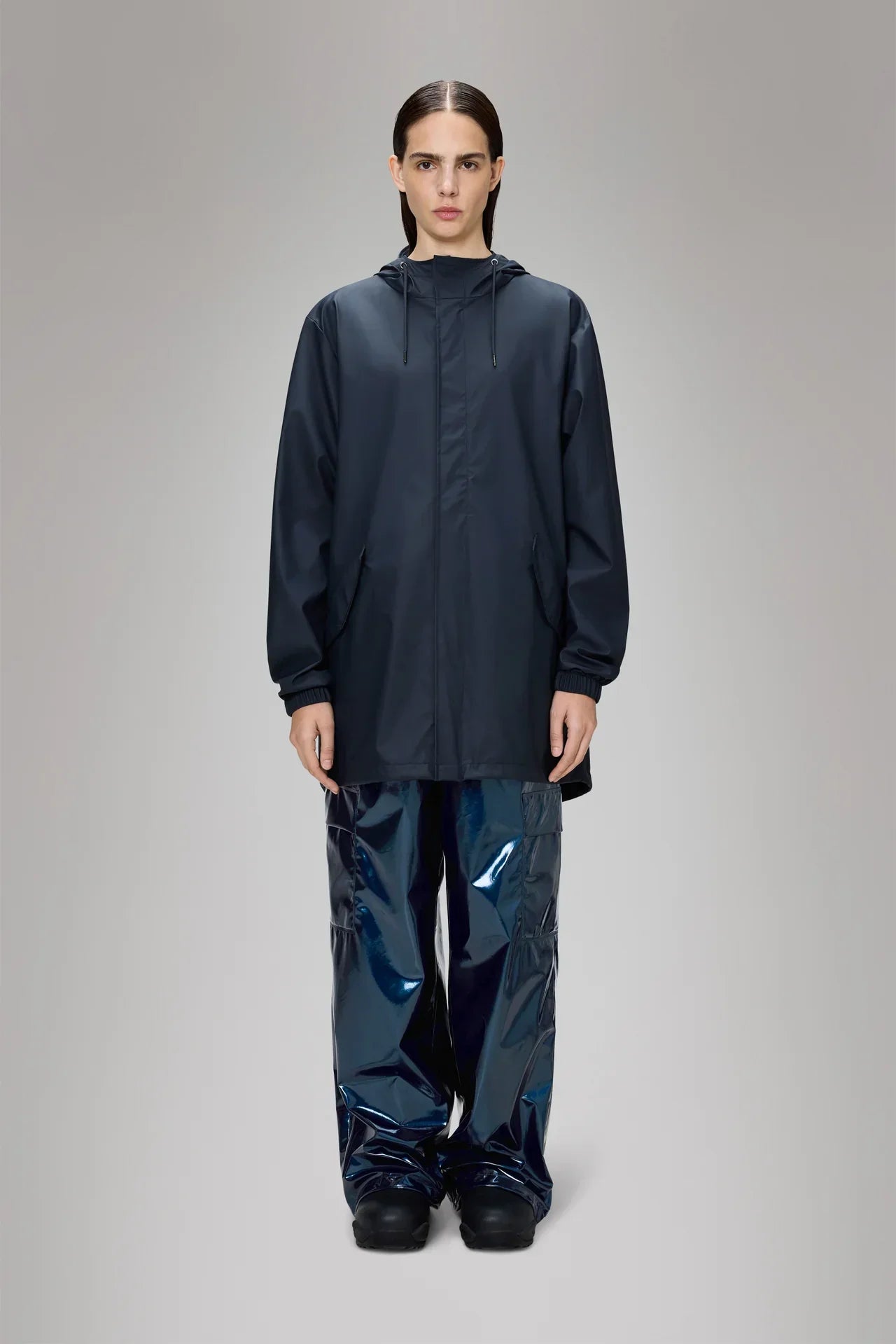 Modella indossa giacca impermeabile blu con cappuccio e pantaloni over lucidi, sfondo neutro.