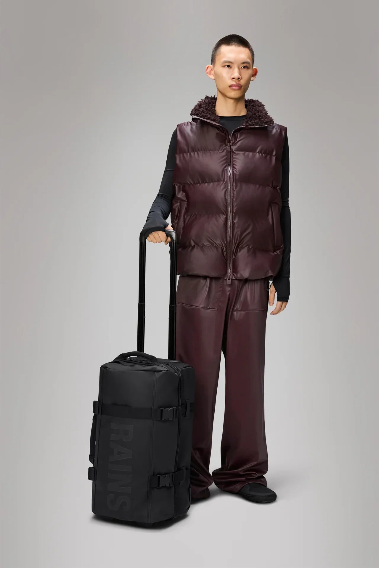 Modello con gilet imbottito marrone scuro e trolley nero da cabina con manico estensibile.