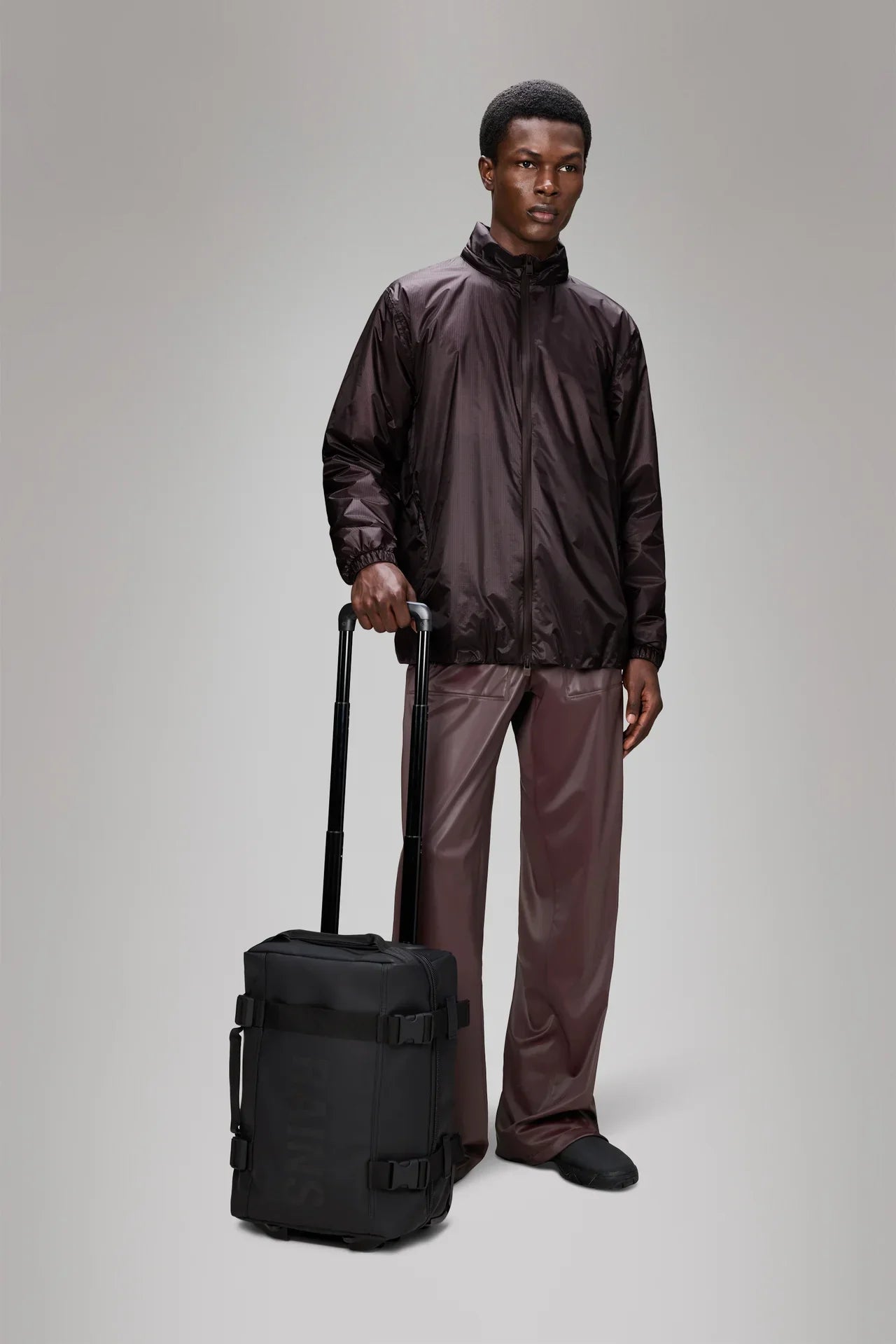 Uomo con giacca tecnica marrone e pantaloni lucidi, trolley nero da cabina moderno