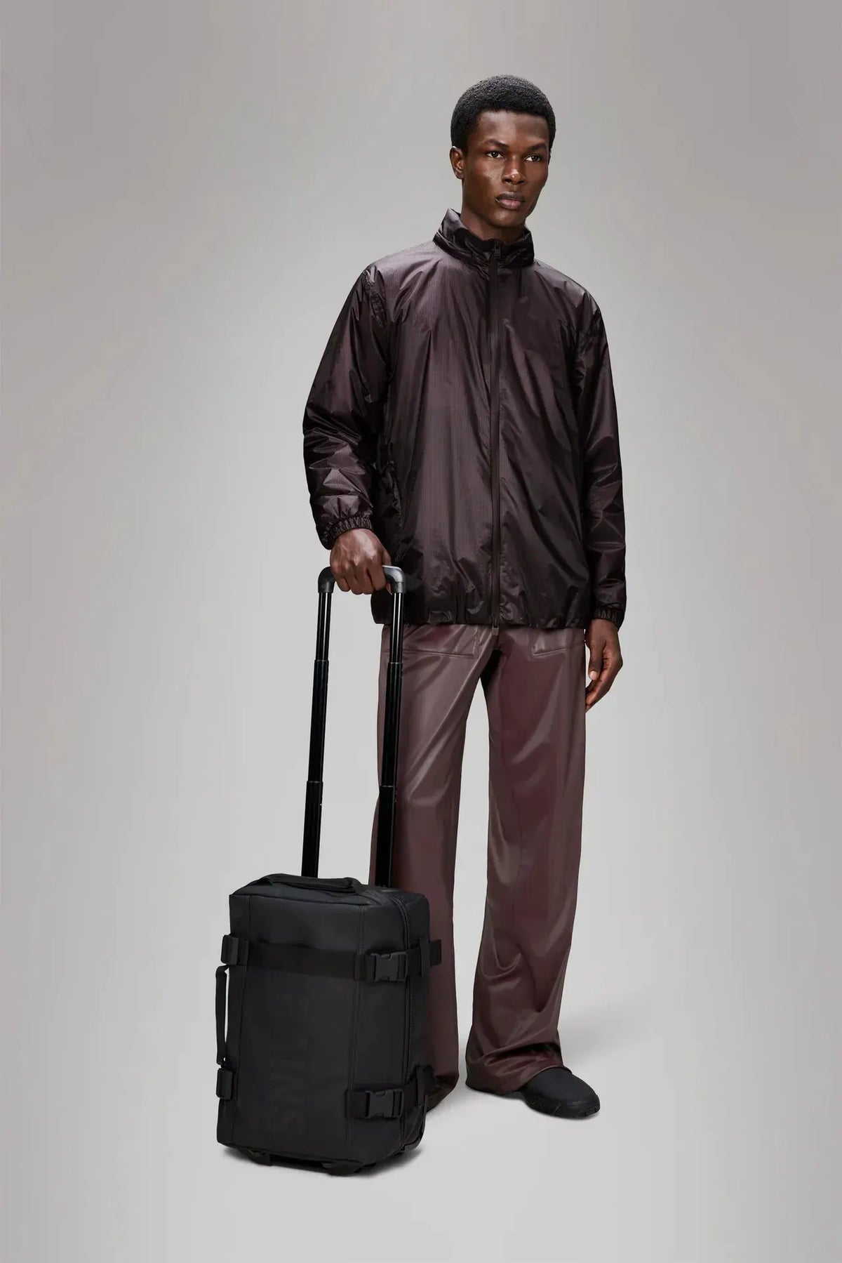Uomo con giacca tecnica marrone e pantaloni lucidi, trolley nero da cabina moderno