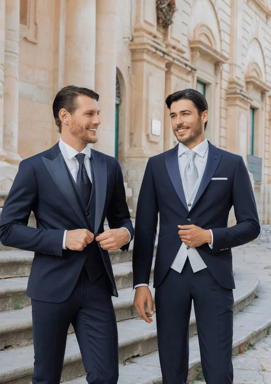 Due uomini indossano eleganti abiti da cerimonia blu, sorridenti all'esterno di un edificio storico.