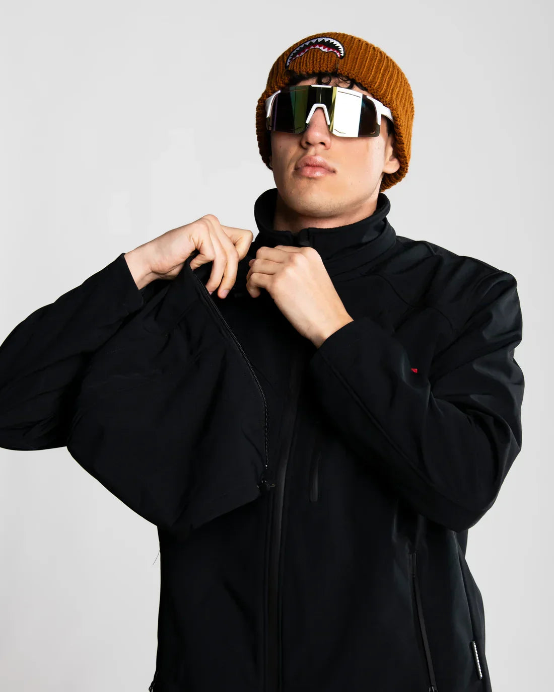 Giovane con giacca nera softshell, cappello marrone e occhiali da sole moda, sfondo neutro.