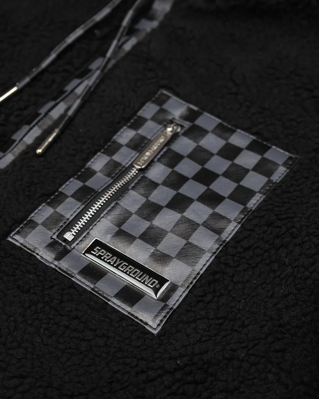 Dettaglio tasca con zip su giacca in pelliccia sintetica nera con pattern a scacchi grigi.