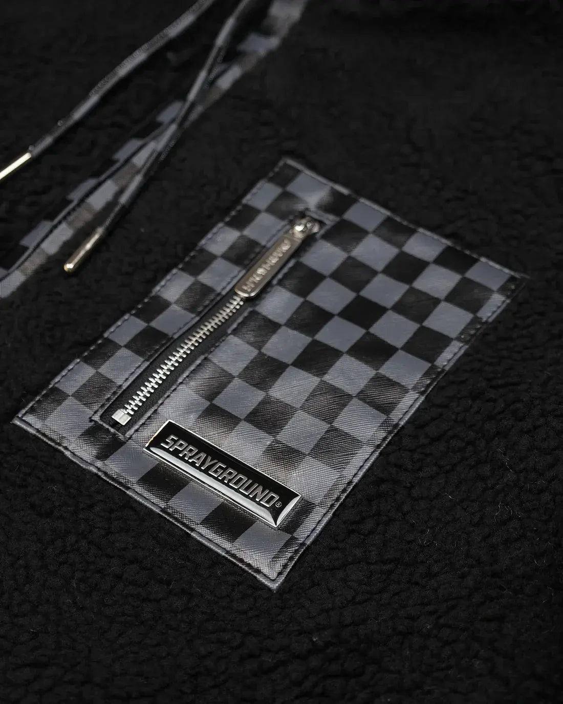 Dettaglio tasca con zip su giacca in pelliccia sintetica nera con pattern a scacchi grigi.