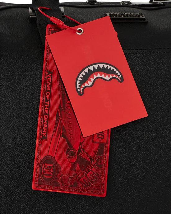 Dettaglio etichette rosse su borsa nera in pelle con motivo bocca di squalo.