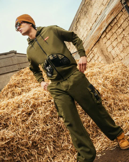 Uomo con felpa e pantaloni verdi, cappuccio, marsupio e berretto, posa outdoor su paglia