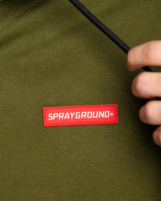 Dettaglio felpa verde con etichetta SPRAYGROUND rossa, mano che tiene il cordino.