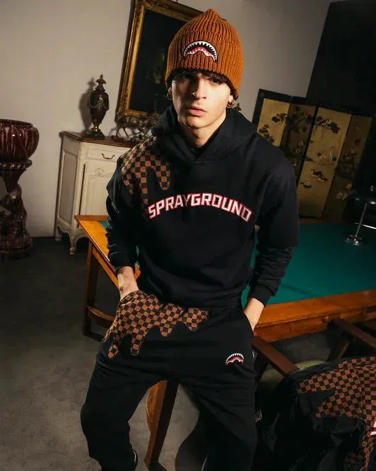 Uomo con felpa nera Sprayground, motivo a scacchi marroni e cappello marrone, moda streetwear.