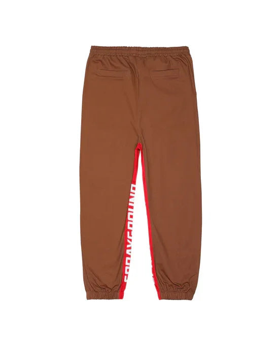 Pantaloni jogger marroni con tasche posteriori, banda rossa laterale e scritta bianca.