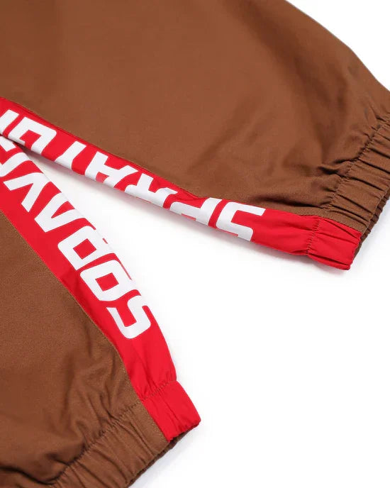Dettaglio pantaloni sportivi marroni con inserti rossi e scritta bianca, elastico alle caviglie.