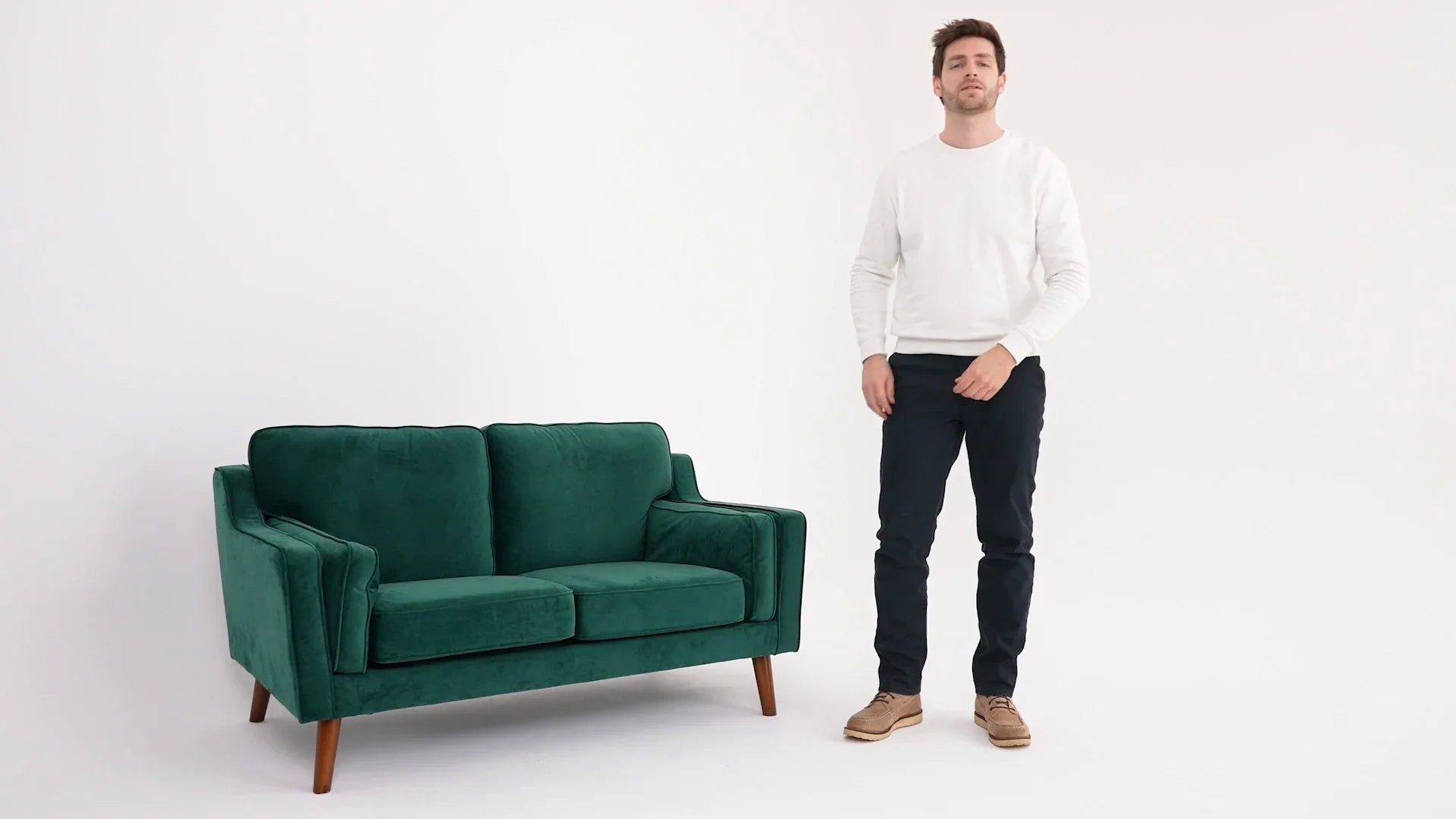 Uomo con maglione bianco, pantaloni scuri e scarpe beige accanto a divano verde elegante.