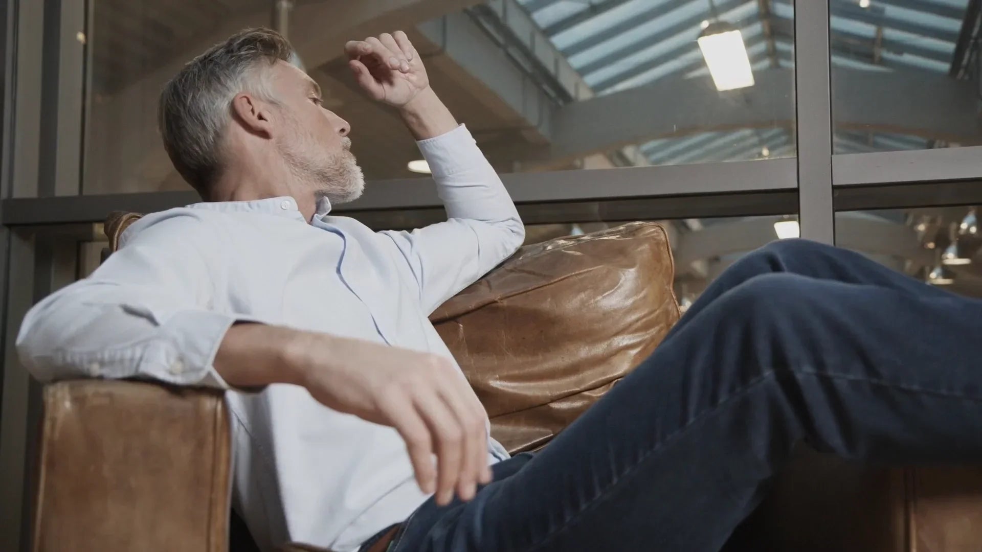 Uomo maturo elegante in camicia bianca e jeans seduto su poltrona in ambiente moderno